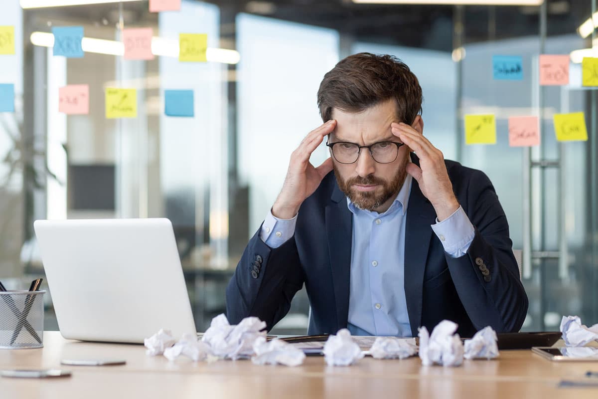 businessman sitting at desk with laptop and crumpled papers in front of him struggling to think of new ideas