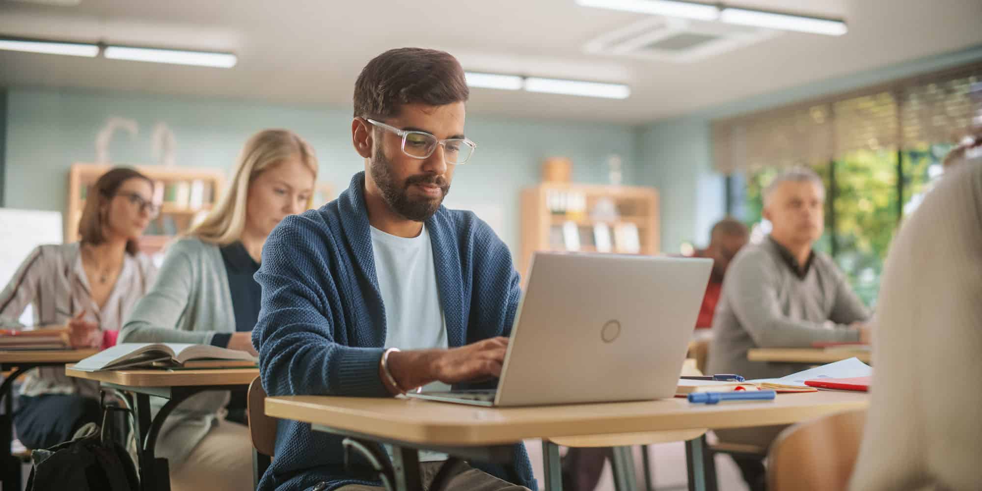 adult students sit at desks in classroom using education software on laptops