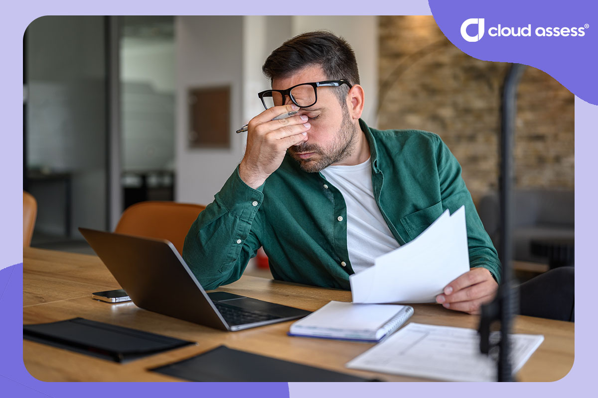 employee holds head in frustration while using laptop and reviewing paper documents