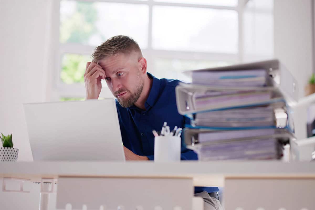 stressed business man looks at laptop holding head with pile of papers on his desk indicating threat of stress and burnout
