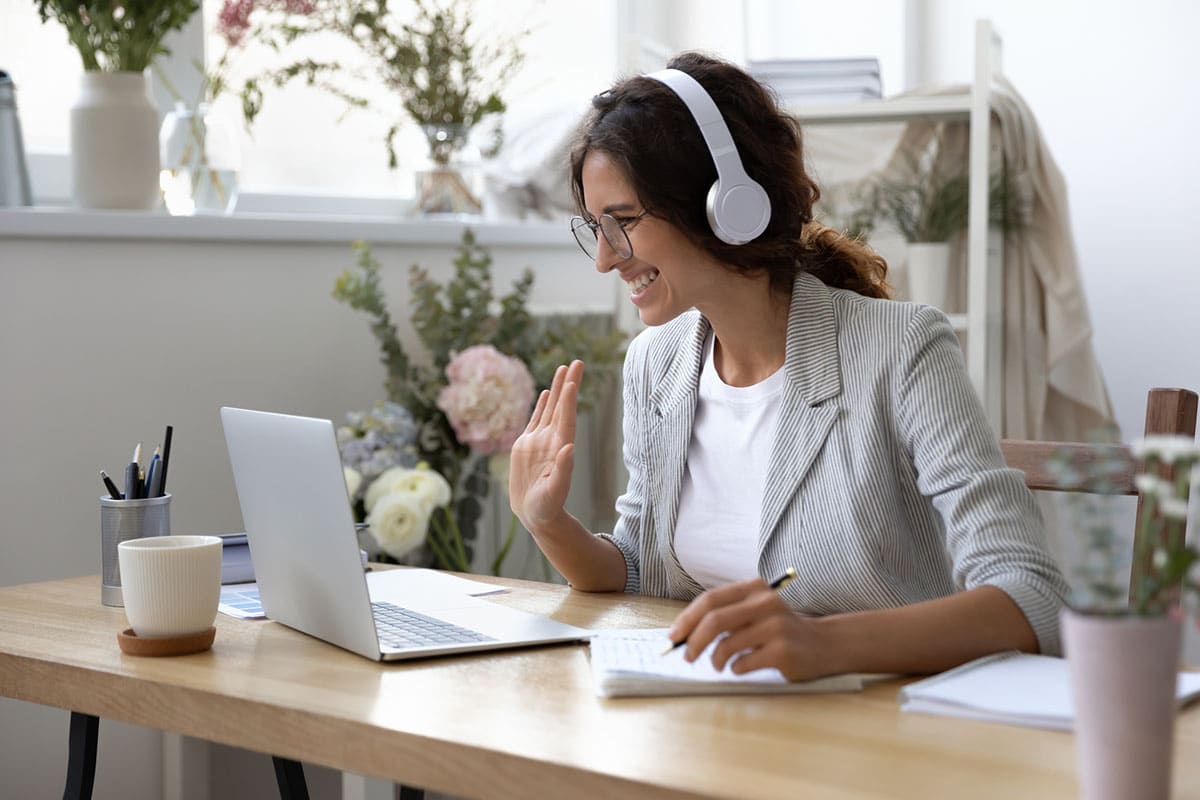 woman with headphones sits at desk and waves at laptop screen during induction training