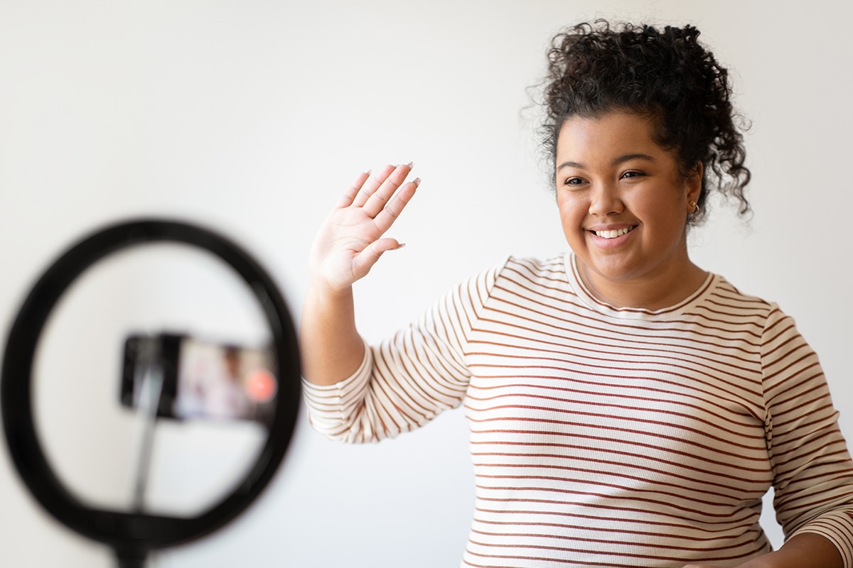 young woman waves as camera while filming video on phone